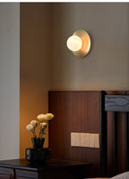 The Clérachic Orb sconce glowing above a walnut headboard, ranunculus in a vase below.