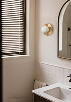 Clérachic Orb sconce beside an arched mirror in a bathroom, daylight through timber blinds.