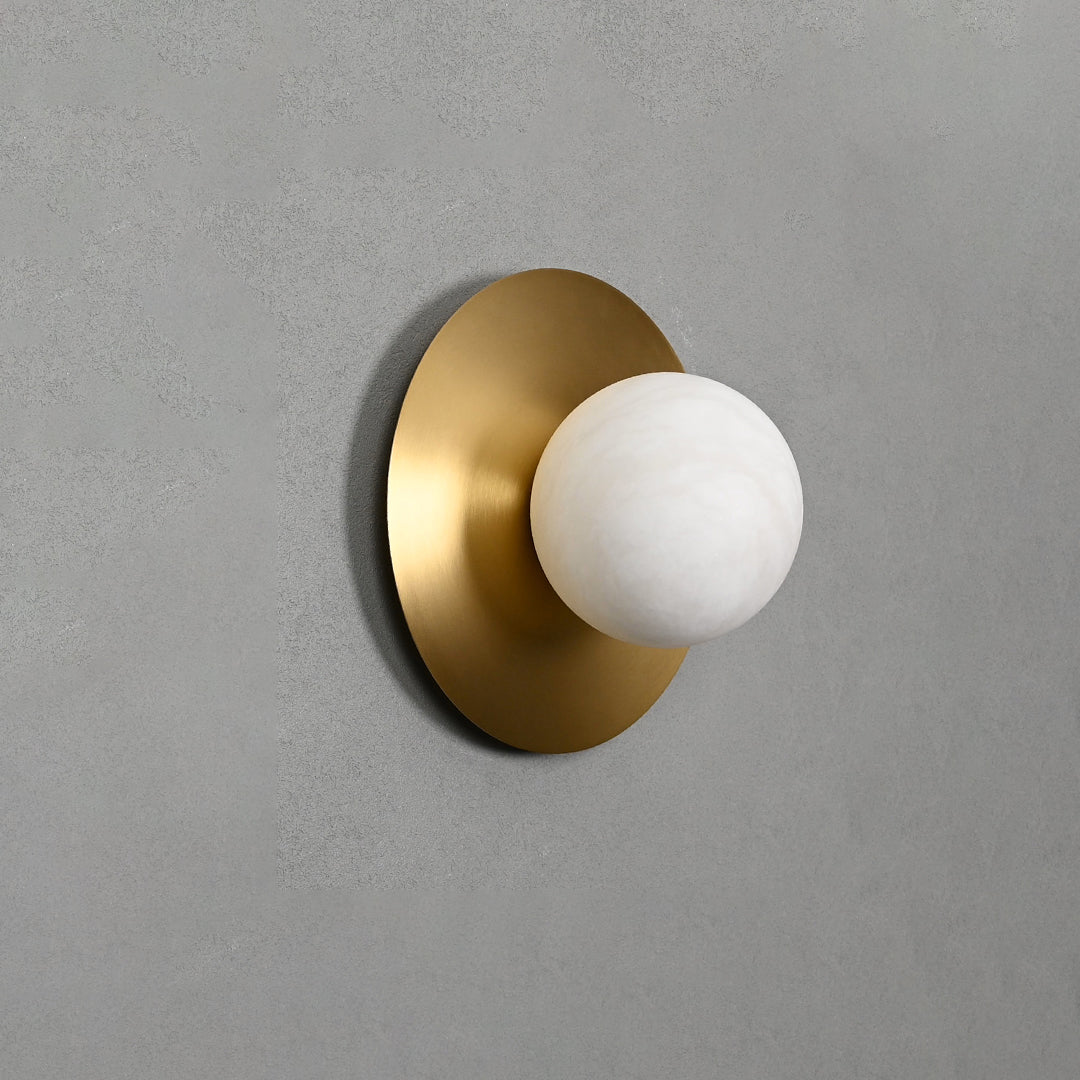 The Clérachic Orb sconce unlit on grey plaster, brass disc catching a soft sheen of daylight.