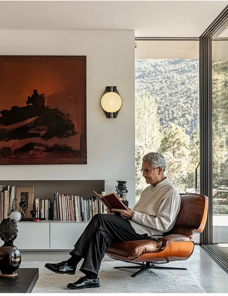 Clérachic Orb sconce on a living-room wall beside tall glazing and a leather lounge chair.