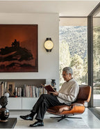 Clérachic Orb sconce on a living-room wall beside tall glazing and a leather lounge chair.
