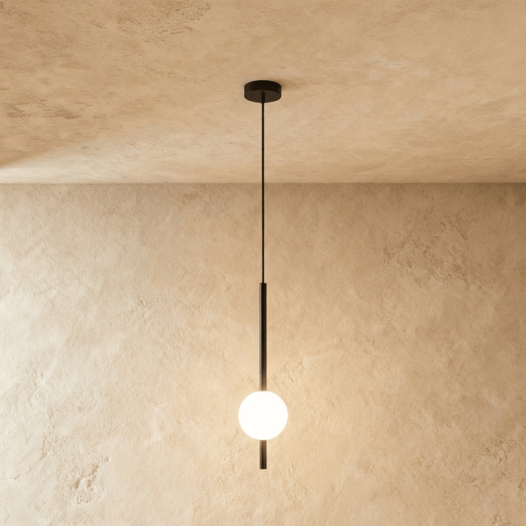 Clérachic single-ball orb pendant light in a matte black finish, glowing warm against a lime-plaster wall backdrop.