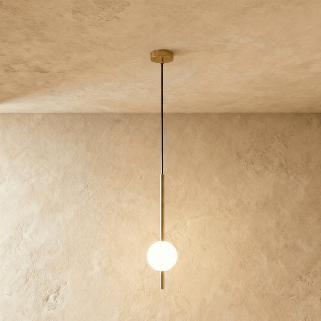 Clérachic single-ball orb pendant light in a brushed brass finish, glowing warm against a lime-plaster wall.