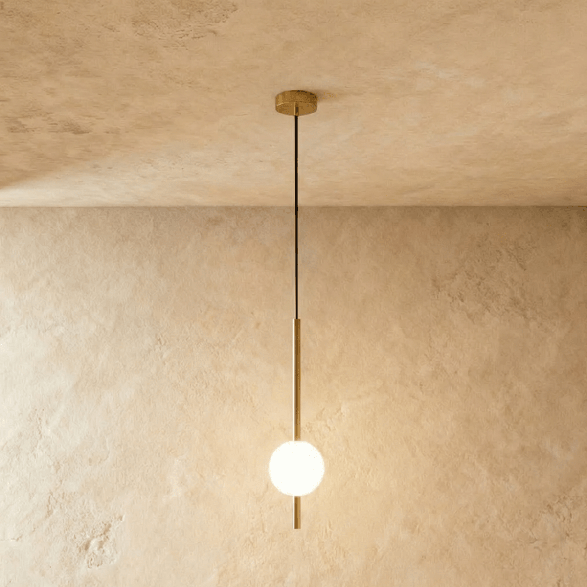 Clérachic single-ball orb pendant light in a brushed brass finish, glowing warm against a lime-plaster wall.