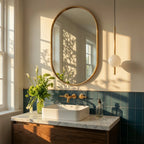 Clérachic single-ball orb pendant light in brushed brass hung beside an oval mirror in a sunlit blue-tiled bathroom.