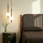 Clérachic twin-head orb pendant light in matte black glowing warm beside an antique wood headboard and nightstand.