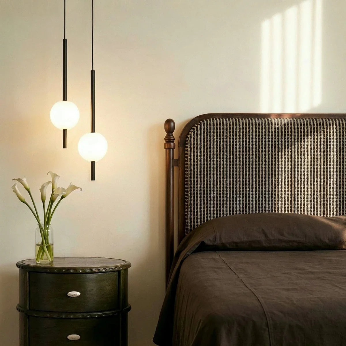Clérachic twin-head orb pendant light in matte black glowing warm beside an antique wood headboard and nightstand.