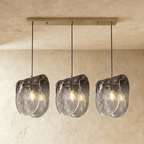 Clérachic three-head linear pendant lit warm, rippled smoke-grey glass shades under a long gold-tone canopy bar.