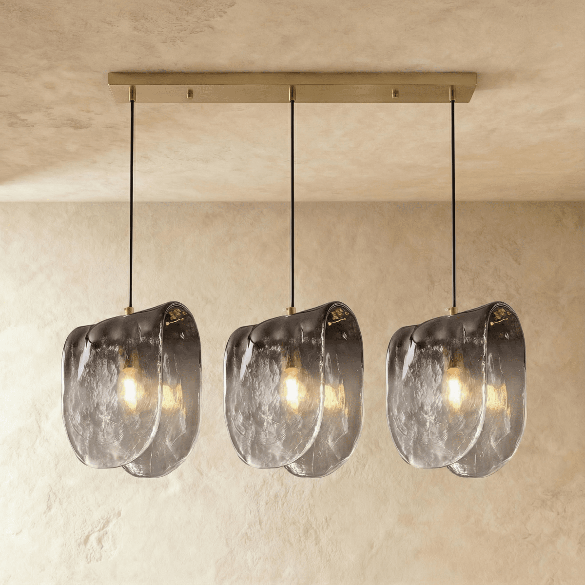 Clérachic three-head linear pendant lit warm, rippled smoke-grey glass shades under a long gold-tone canopy bar.