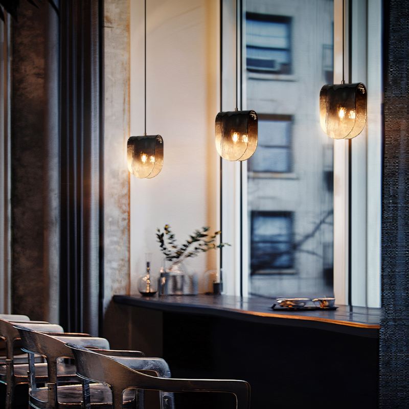 Clérachic smoke-grey glass pendants glowing at different heights over a dark counter, with stools and tall windows.