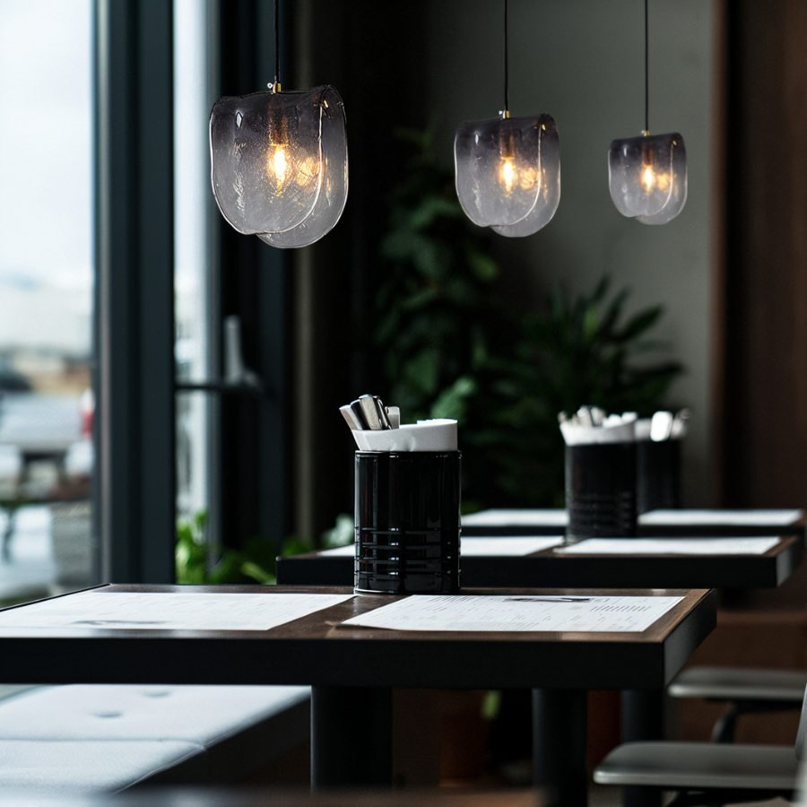 Clérachic smoke-grey glass pendants glowing warm over café tables, with a plant and tall windows behind.