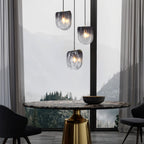 Three Clérachic smoke-grey glass pendants at staggered heights over a round dining table, with a misty forest view.