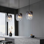 Three Clérachic smoke-grey glass pendants lit warm over a stone kitchen island, with a vase and bowl beside a window.