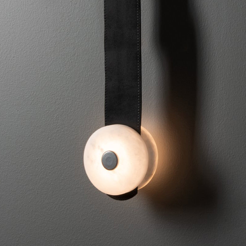 Black leather strap and one glowing alabaster disc of the Clérachic Orinos, seen close beside a shadowed wall.