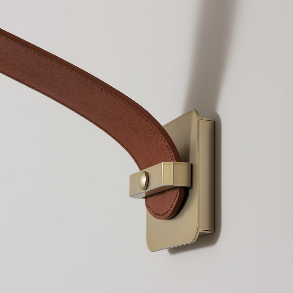 Detail of the Clérachic Orinos wall bracket, brushed brass with the stitched cognac strap looped through it.