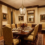 Clérachic brass lantern pendant above a long polished dining table, with gold curtains and gilt-framed portraits behind it.