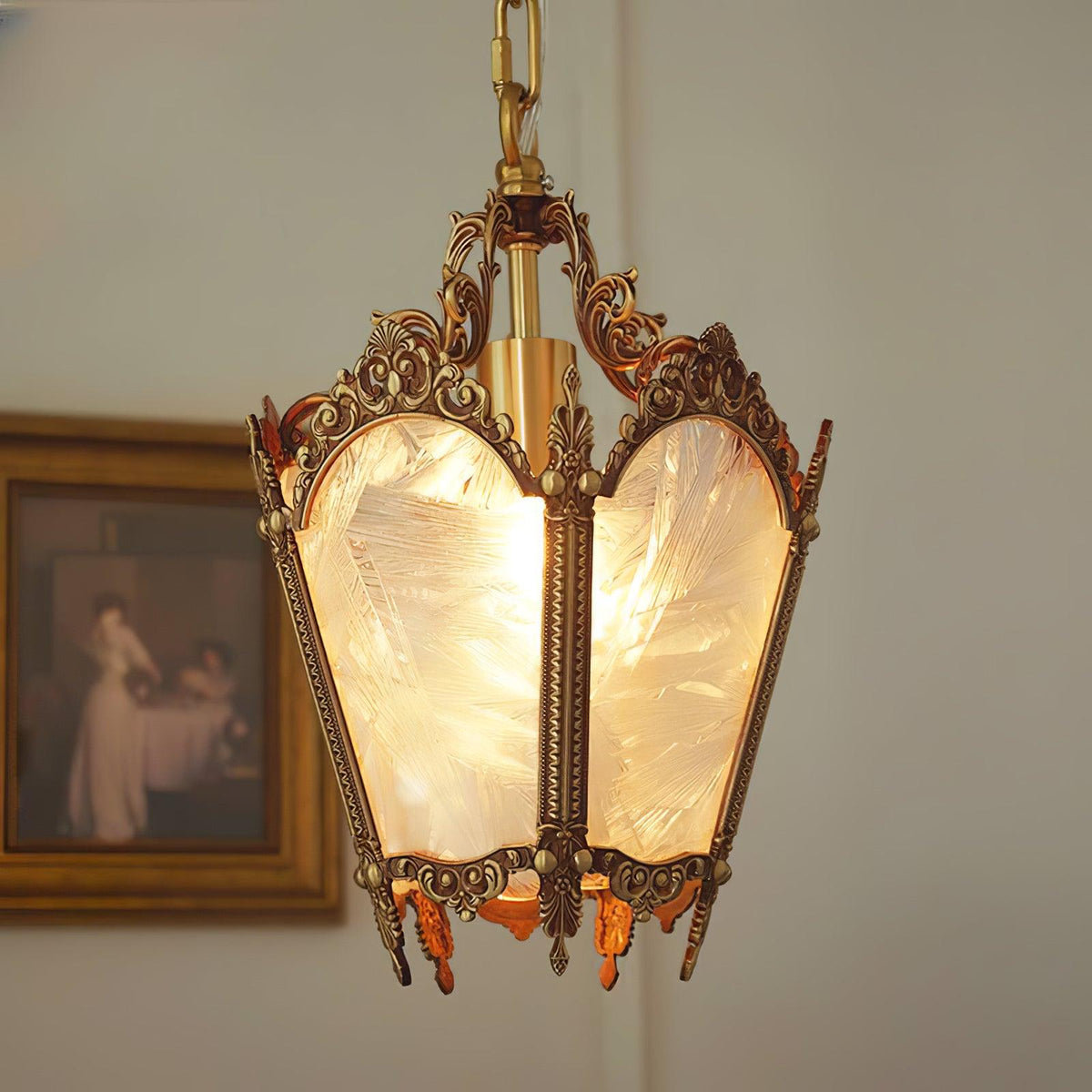 Lit brass lantern pendant with ornate scrollwork and frosted panes, hung beside a framed painting, from Clérachic.