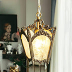 Lit Clérachic lantern pendant with a darker aged-brass frame, hung near a window with sheer curtains and a framed portrait.