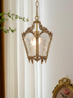 Clérachic brass lantern pendant hung beside white curtains, with leafy stems above and a gilt frame in the corner.