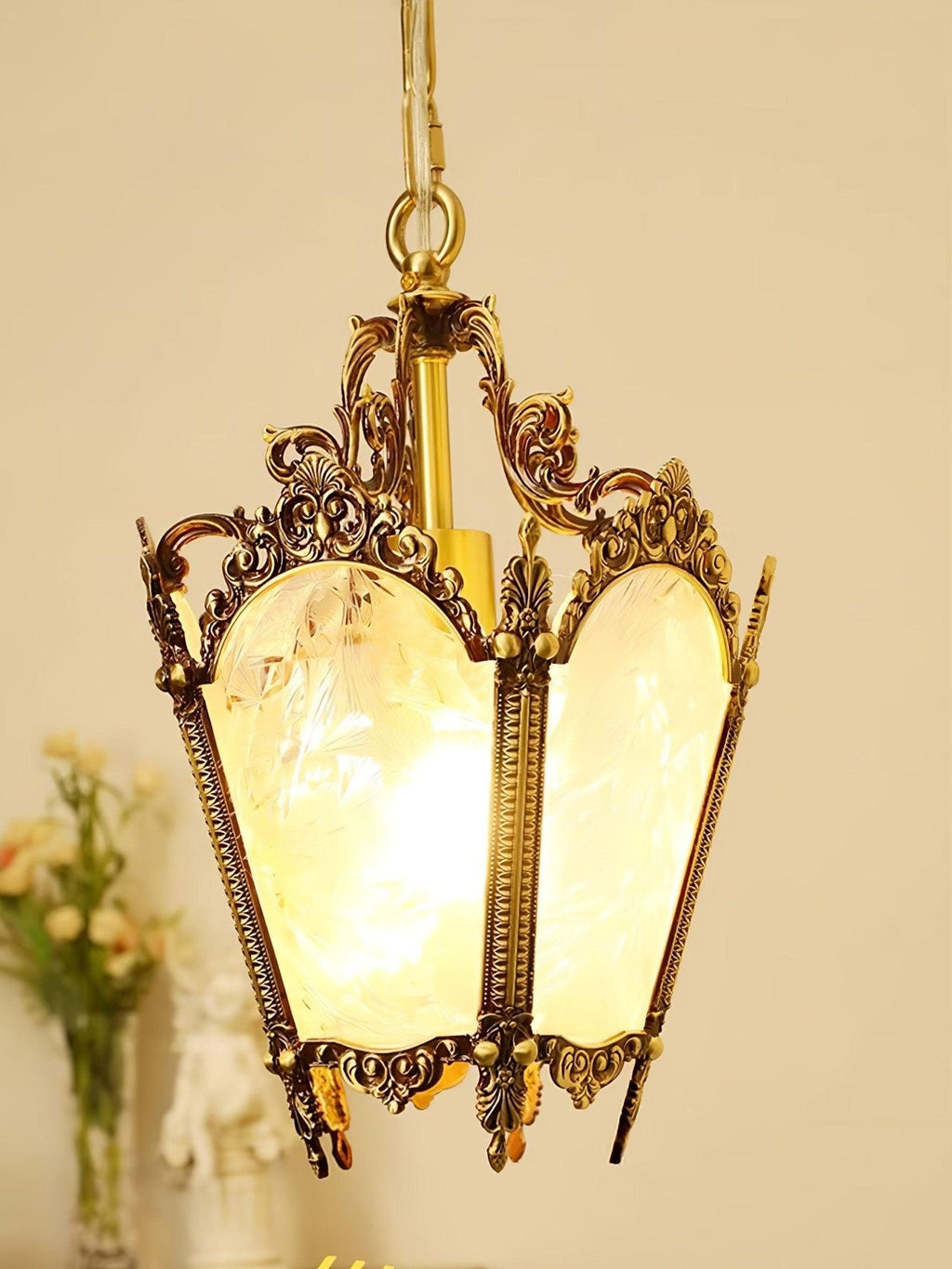 Lit brass lantern with scrolled arms and warm glowing panes, a vase of pale flowers behind it, from Clérachic.