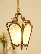 Lit brass lantern with scrolled arms and warm glowing panes, a vase of pale flowers behind it, from Clérachic.