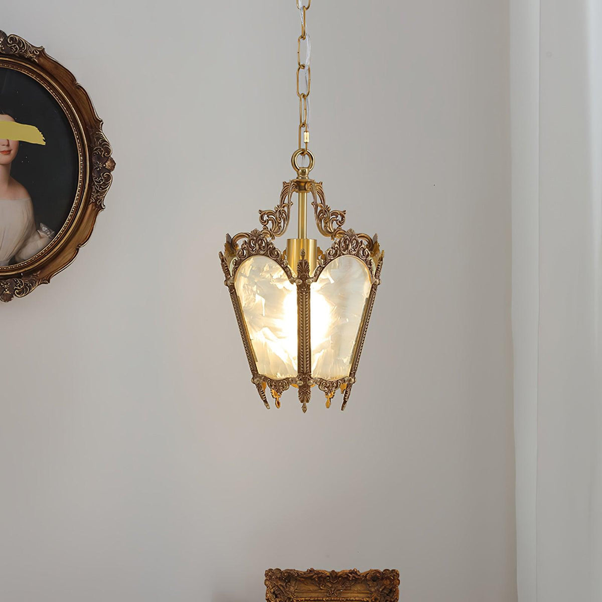 Brass lantern pendant on a chain against a white curtain and wall, with an oval portrait at the left, from Clérachic.