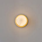 Clérachic Orrila wall light lit head-on, a ring of brass framing the alabaster face.