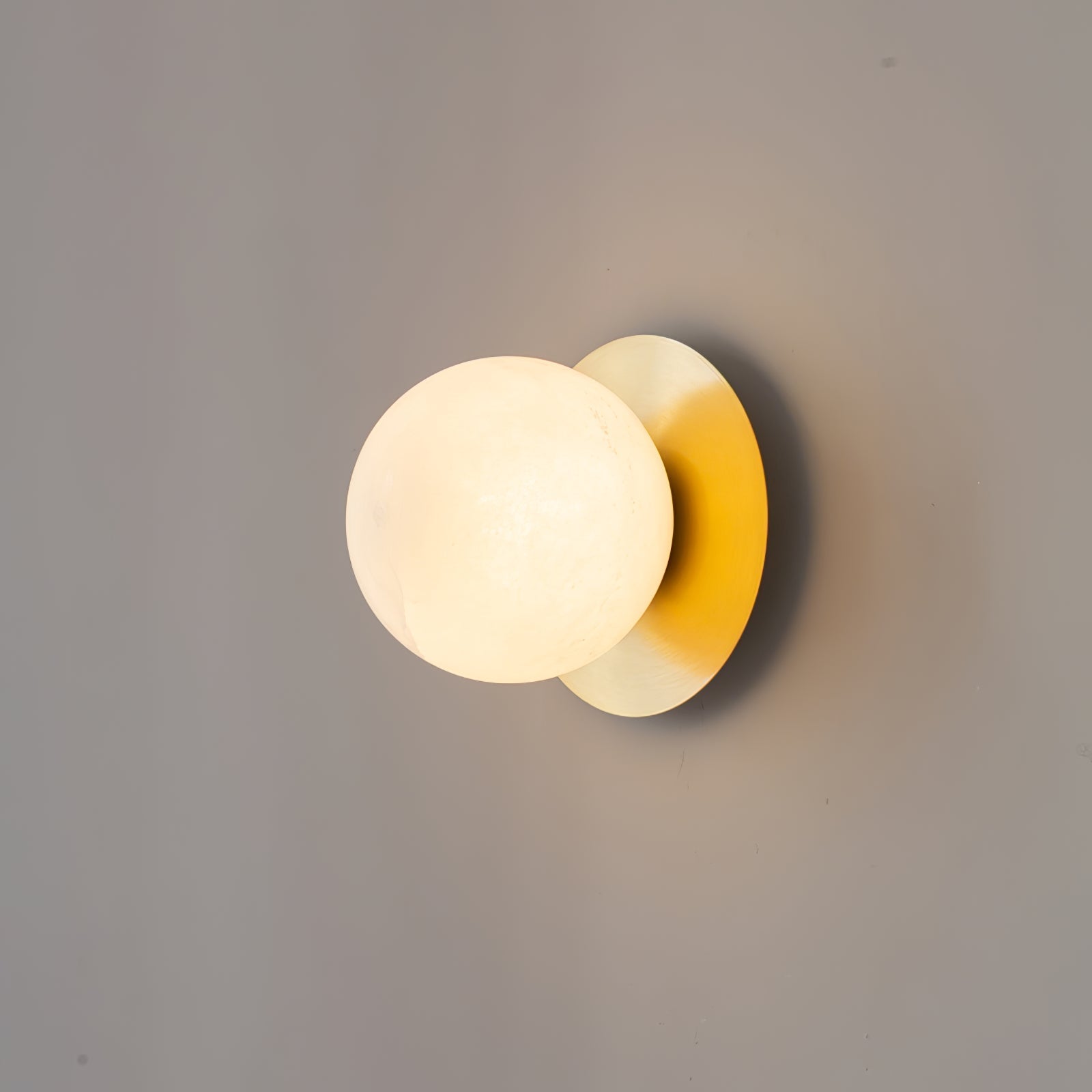 Clérachic Orrila wall sconce lit on grey, a soft shadow cast behind the brass plate.