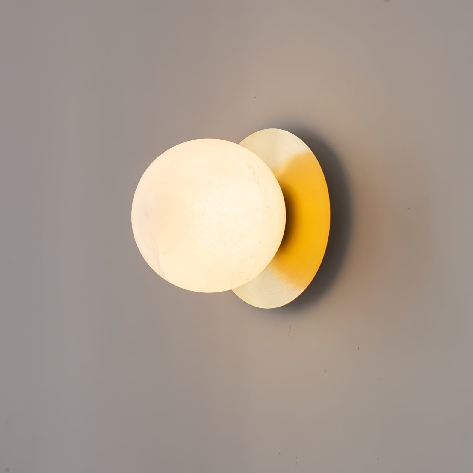 The Clérachic Orrila sconce angled on a grey wall, globe glowing beside the brass disc.