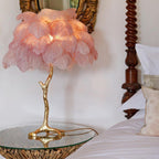 Close view of this Clérachic feather lamp glowing pink on a glass-topped side table beside a carved four-poster bed and a gilt mirror.