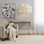A cream-feathered Clérachic table lamp on a silver stem, on a slim console beside a beige armchair with a white throw.