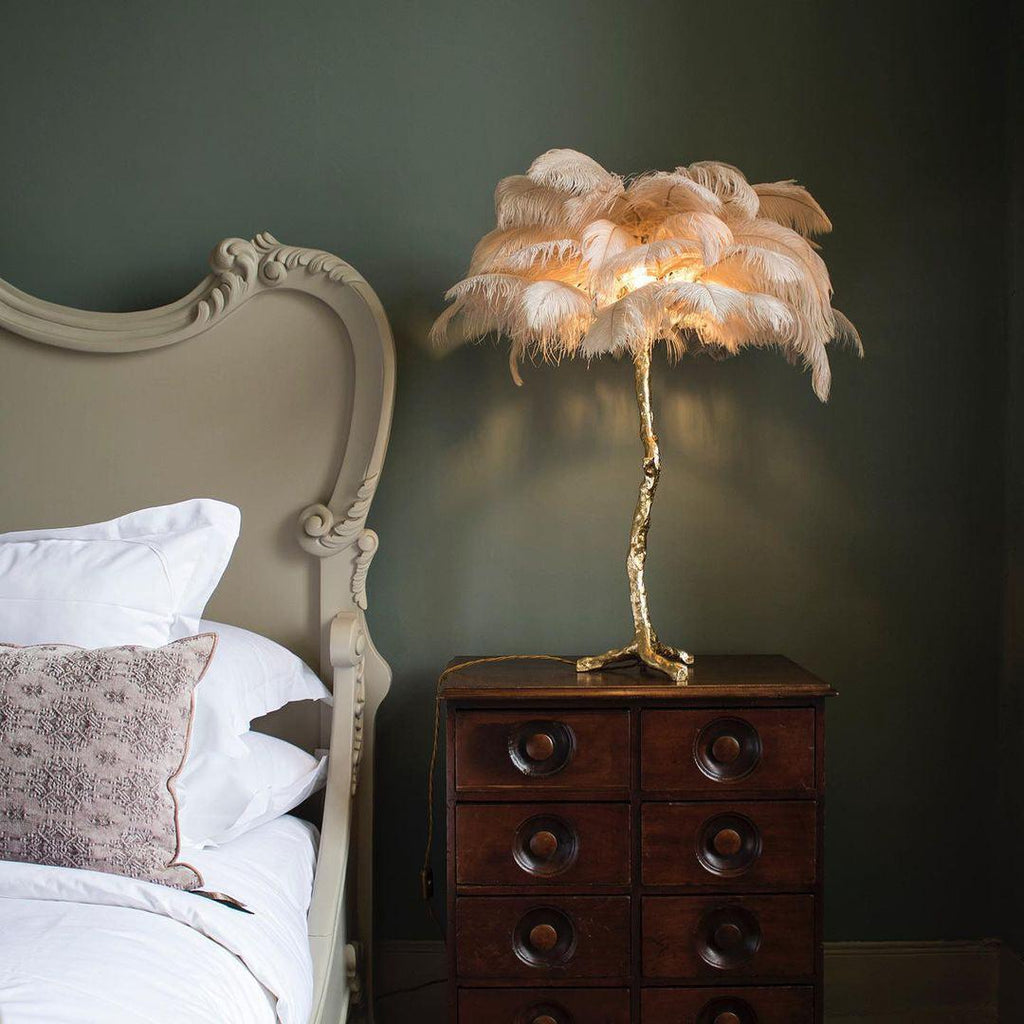 A Clérachic table lamp with soft peach feathers lit up on a brass branch stem, on a dark chest of drawers beside a cream bed.