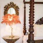 Clérachic feather lamp with orange plumes glowing on a glass table, with a spiral-carved wooden bedpost and gilt mirror behind.