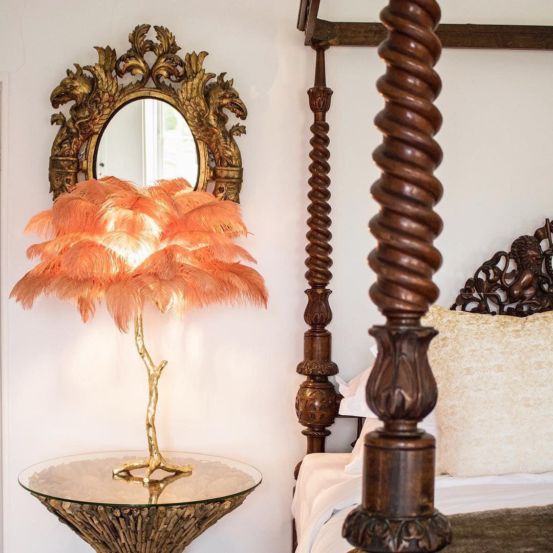 Clérachic feather lamp with orange plumes glowing on a glass table, with a spiral-carved wooden bedpost and gilt mirror behind.