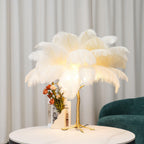 A white Clérachic feather table lamp lit on a round marble-topped table, with a vase of orange flowers and a green velvet chair behind.