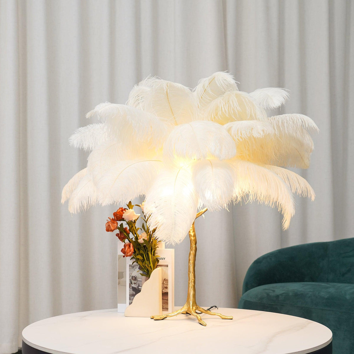 A white Clérachic feather table lamp lit on a round marble-topped table, with a vase of orange flowers and a green velvet chair behind.