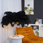 Black-feathered Clérachic table lamp with a brass branch stem on a white fluted side table, beside a mustard velvet lounge chair.
