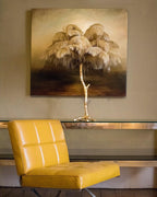 Clérachic feather table lamp with tan plumes in front of an oil painting, on a glass shelf beside a yellow leather chair.