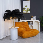 Clérachic black feather table lamp lit on a white fluted side table beside a mustard lounge chair, with a framed plant print above.