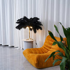 A lit Clérachic black-feathered table lamp on a fluted side table, with white curtains behind and large green leaves in the corner.