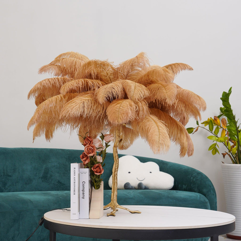 Clérachic table lamp with camel-coloured feathers on a round table with a vase of roses and books, in front of a green velvet sofa.