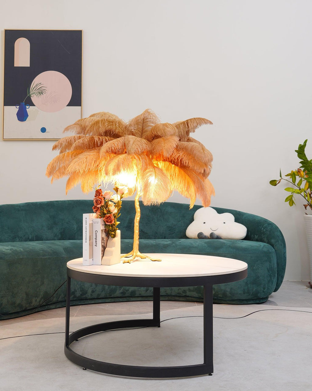Lit Clérachic feather lamp with tan-gold plumes on a black-framed coffee table, in a room with a green sofa and a cloud-shaped cushion.