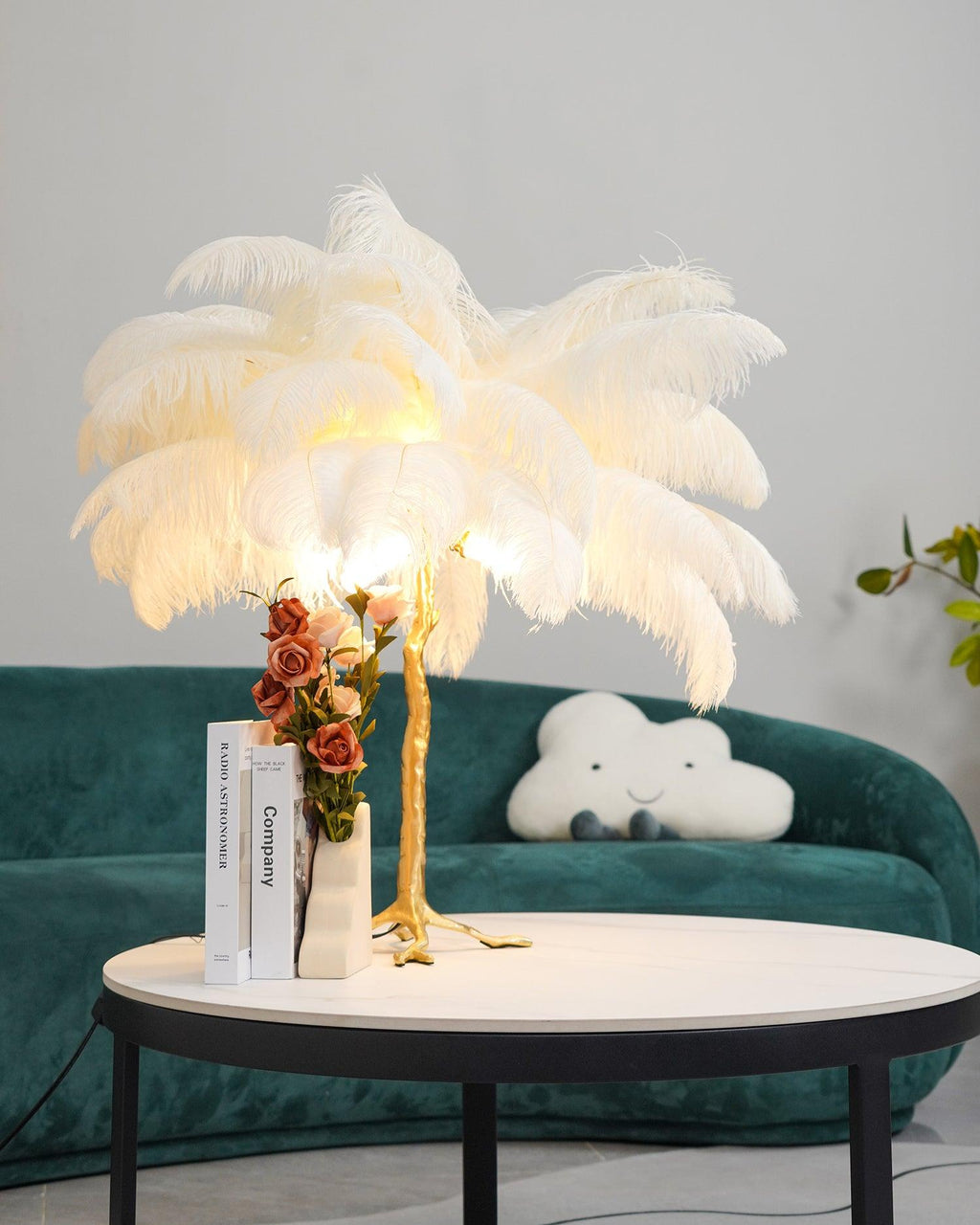 A white-feathered Clérachic table lamp glowing on a marble-topped table, with roses, books and a green sofa behind it.
