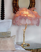Clérachic feather lamp with soft pink plumes lit up beside white pillows, with a carved bedpost and a round gilt mirror behind.
