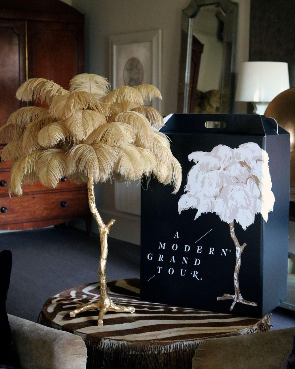 A Clérachic table lamp with sand-coloured plumes on a zebra-print stool, with a black gift bag and a lamp beside it.