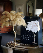 A Clérachic table lamp with sand-coloured plumes on a zebra-print stool, with a black gift bag and a lamp beside it.