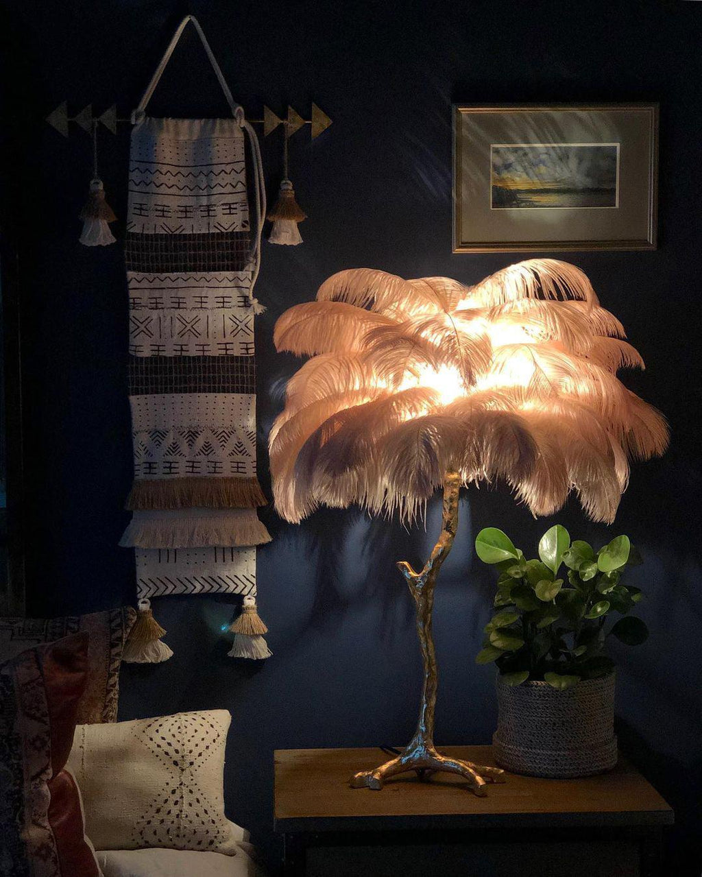 Lit Clérachic feather table lamp glowing peach in a dark blue room, on a wooden bedside table beside a small green plant.