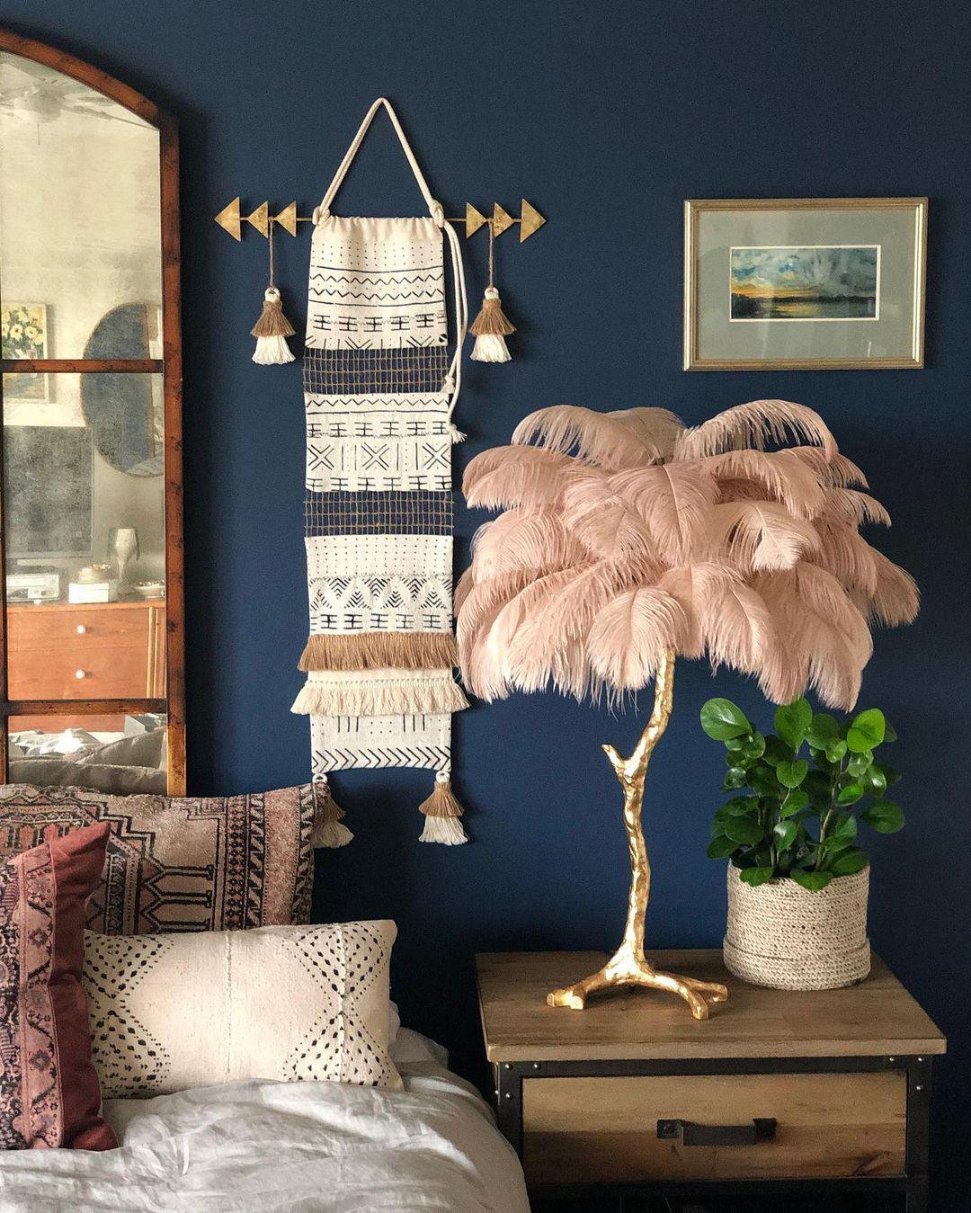 Clérachic table lamp with pale pink feathers on a wooden bedside table, against a navy wall with a woven wall hanging.