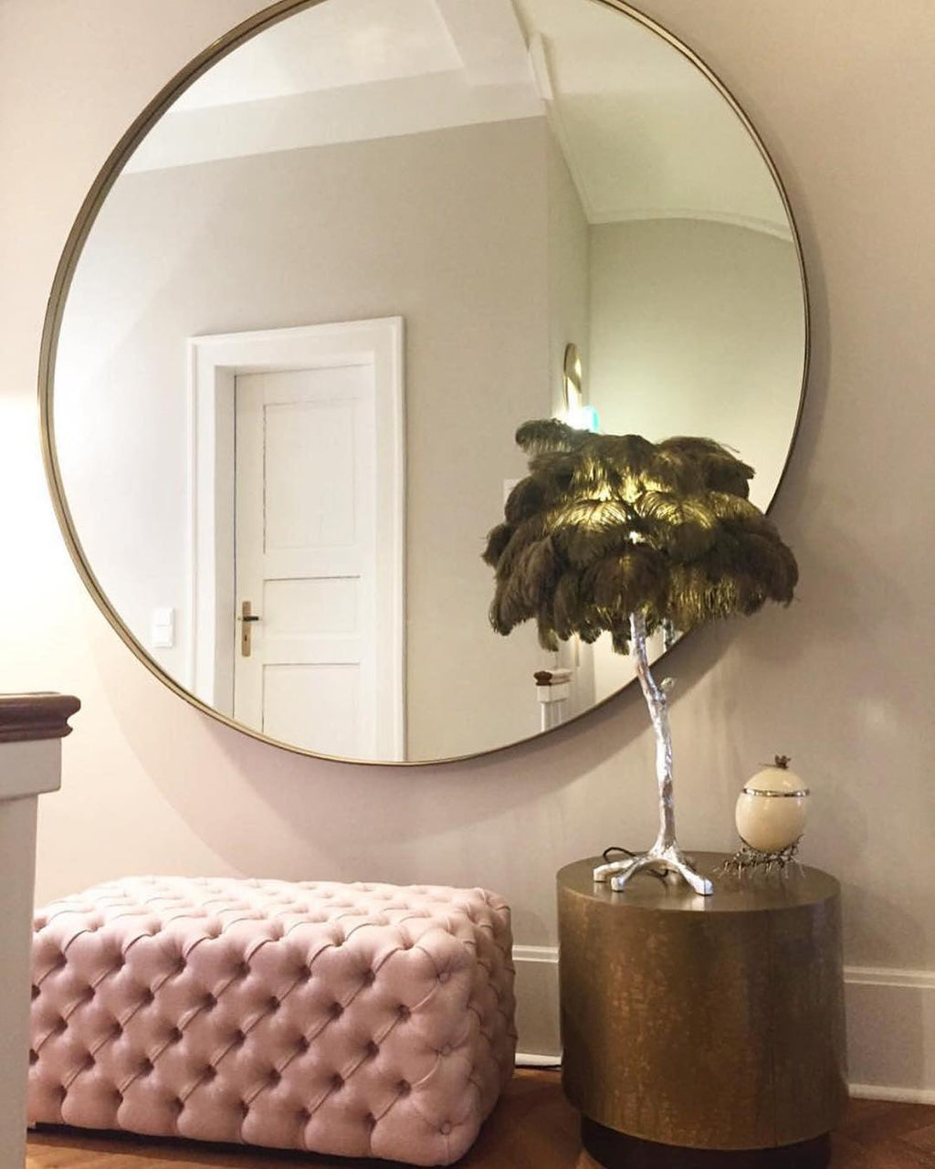 A dark-feathered Clérachic table lamp on a silver stem on a bronze side table, beside a pink tufted ottoman and a large round mirror.