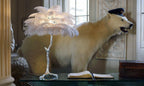 Clérachic table lamp with white feathers on a silver stem on a glass desk, in front of a stuffed polar bear wearing a cap.